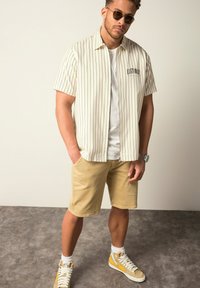 Camicia a maniche corte a righe color crema con righe nere, abbinata a pantaloni corti khaki. Indossata con sneakers con dettagli gialli. Look casual.