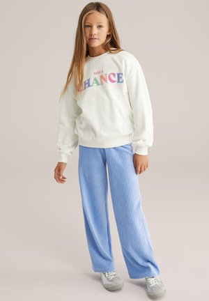 Bela majica z napisom "TAKE A CHANCE" v večbarvnih črkah, kombinirana z modrimi širokimi hlačami z rebrastim vzorcem in sivimi supergami.