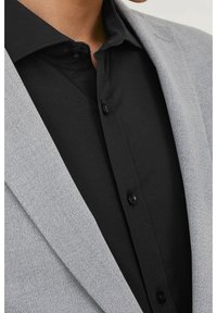 Grauer Blazer mit strukturierter Oberfläche, getragen über einem schwarzen Hemd mit Knopfleiste und klassischem Kragen.