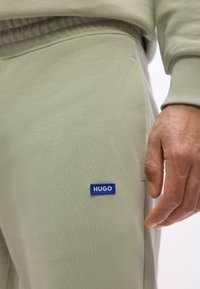 Ανοιχτό πράσινο παντελόνι φόρμας με ελαστική μέση και μπλε ορθογώνιο λογότυπο "HUGO" κοντά στην τσέπη, δίπλα σε ένα χαλαρό χέρι ατόμου.