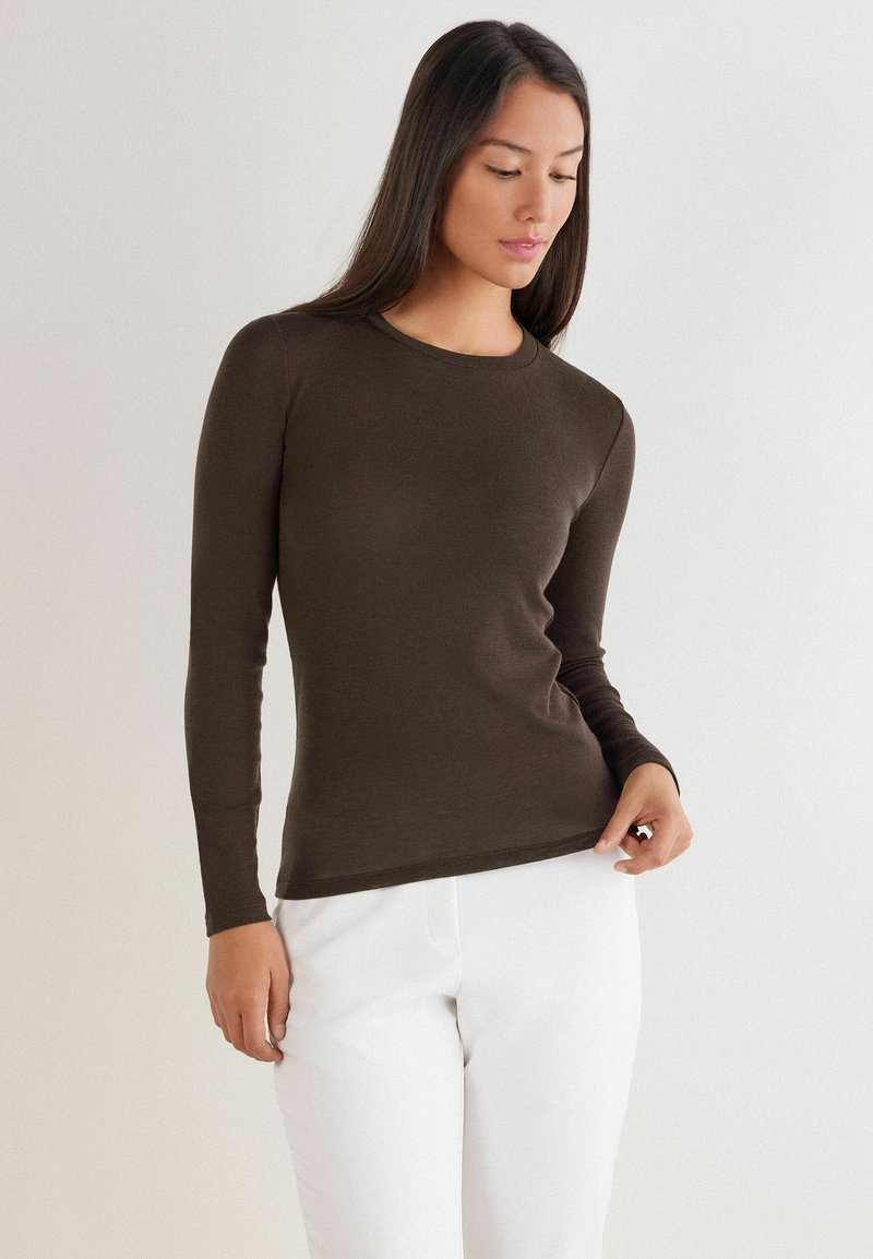 Langärmliges Shirt in Dunkelbraun, aus weichem Material, mit rundem Halsausschnitt und taillierter Silhouette. Kombiniert mit weißen Hosen.