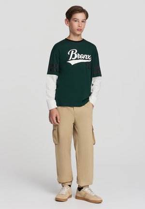 T-shirt verde a maniche corte con la scritta bianca "Bronx" e strisce nere, abbinato a pantaloni cargo beige e sneakers beige.