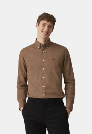 Uomo sorridente che indossa una camicia marrone con bottoni e colletto e pantaloni neri, mani nelle tasche, in piedi contro uno sfondo chiaro uniforme.