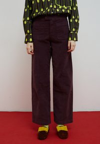 Pantalon à jambes larges de couleur violet foncé en tissu léger, associé à une chemise noire avec des motifs floraux verts et des boutons. Chaussures marron avec des accents jaunes.