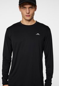 Svart långärmad skjorta i ett mjukt, lätt tyg. Har en liten grå logotyp på bröstet och en rund halsringning.