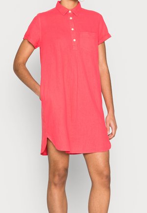 Nainen, joka pukeutunut korallinväriseen lyhythihaiseen polopaitamekkoon, jossa on napit, rintatasku ja sivuhalkiot, seisoo yksiväristä taustaa vasten.