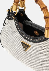 Canvas handbag in cream with black accents, featuring a gold logo, braided bamboo handle, and stitched detailing. Textured fabric design.