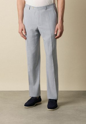 Mann trägt hellblau-weiß gestreifte Hosen mit vertikalen Streifen und marineblaue Schuhe, steht vor einem einfarbigen beigen Hintergrund.