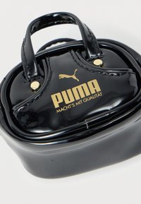 Γυαλιστερή μαύρη τσάντα Puma με χρυσό λογότυπο και γερμανικό κείμενο "Macht's mit Qualität" σε λευκό φόντο.