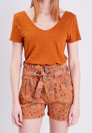 Femme portant un t-shirt à manches courtes couleur orange brûlé avec un col en V et un short taille haute à motif floral orange, avec une ceinture en tissu nouée à la taille.