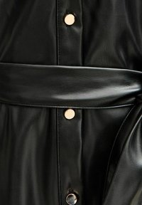 Prenda de cuero negro con una textura suave, diseño estructurado, botones de hardware dorado y un cinturón ancho que acentúa la cintura.