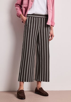 Personne portant un pantalon large noir à rayures blanches verticales, une veste rose boutonnée, un haut blanc et des chaussures marron à enfiler.