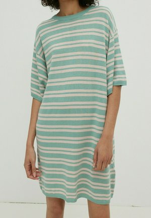 Personne portant une robe ample à manches courtes avec des rayures horizontales blanches et vert clair sur un fond uni.