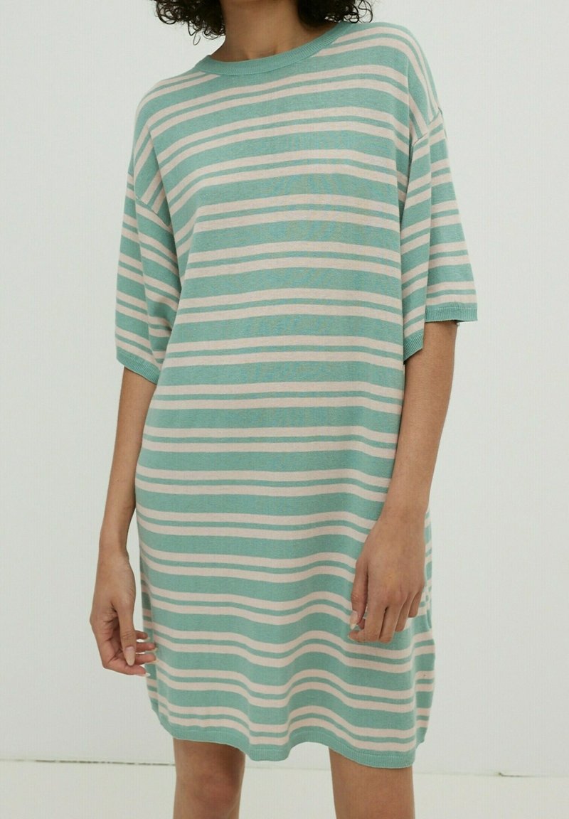 Personne portant une robe ample à manches courtes avec des rayures horizontales blanches et vert clair sur un fond uni.