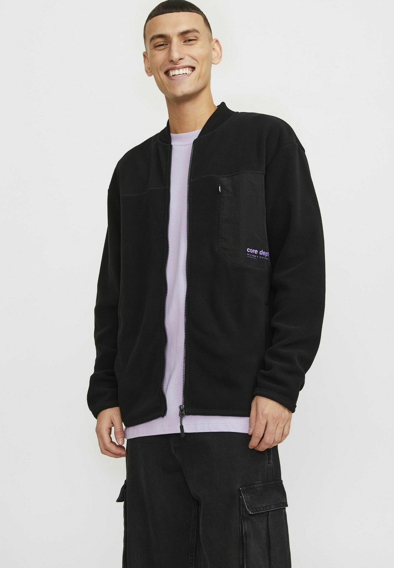 Black fleece jacket with zip front and side pockets; features a purple logo on chest pocket; worn over a light purple shirt.