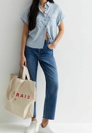 Kvinde i blå stribet kortærmet skjorte, højtaljede blå jeans, hvide sneakers, der holder en beige mulepose med rød tekst, stående op ad en hvid væg.