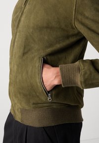 Giacca bomber in suede verde oliva con tasca con zip e polsini a costine. Texture morbida con vestibilità rilassata ed elementi di design classici.