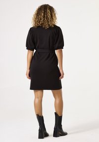 Vestido negro con mangas abullonadas cortas, cintura ceñida con cinturón y un diseño minimalista. Llevado con botas negras de tacón a la altura del tobillo. Fondo liso.