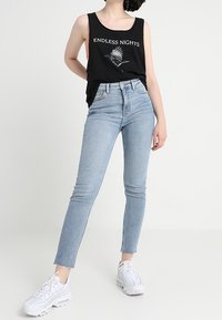 Mujer vistiendo jeans skinny de color azul claro, zapatillas blancas y una blusa sin mangas negra con el texto "NOCHES SIN FIN" y un gráfico de un pájaro.