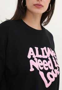 Sweatshirt noir avec le texte brodé en rose "Tout ce dont nous avons besoin, c'est d'amour", col côtelé et coupe décontractée. Texture de tissu douce.