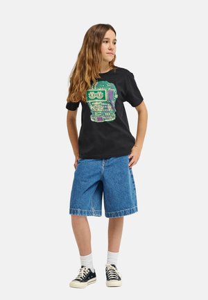 Czarny t-shirt z kolorową grafiką robota, zestawiony z jeansowymi szortami oraz czarno-białymi trampkami. Długie, falujące brązowe włosy.