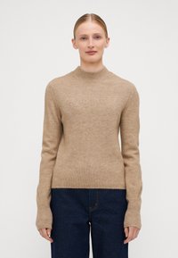 Beige ribbestrikket genser med høy hals og lange ermer. Stoffet er mykt med en jevn tekstur, kombinert med mørkeblå vide jeans.