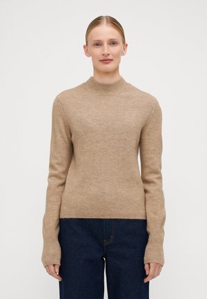 Beige ribbestrikket genser med høy hals og lange ermer. Stoffet er mykt med en jevn tekstur, kombinert med mørkeblå vide jeans.