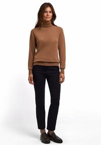 Beige Rollkragenpullover aus weichem Material, kombiniert mit enganliegenden schwarzen Hosen. Auffällige gerippte Bündchen und Saum, getragen mit dunklen Slippern.