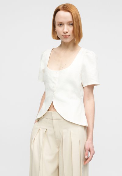 Λευκή μπλούζα από λινό με τετράγωνο ντεκολτέ, κοντά φουσκωτά μανίκια και κλείσιμο με κουμπιά. Συνδυάζεται με μπεζ παντελόνι ψηλόμεσο με πιέτες.