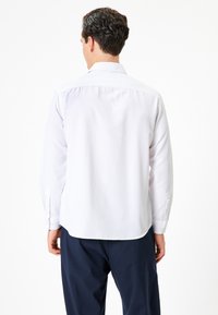 Chemise blanche à manches longues avec un col classique et des poignets à boutons ; présente une texture lisse et un ourlet arrondi. Portée avec un pantalon sombre.