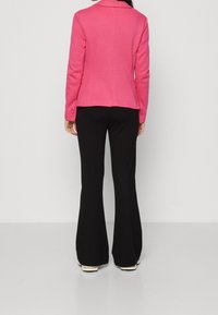 Růžový blejzr s klopovým límcem, dlouhými rukávy a knoflíkovými detaily, v kombinaci s černými zvonovými kalhotami a béžovými botami, viděný ze zad.