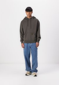 Olive green hoodie with drawstrings and front pocket, paired with wide blue denim jeans and beige sneakers with black soles.