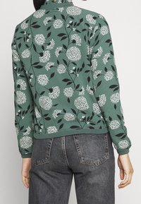 Grüner Pullover mit weißen Blumenmustern und schwarzen Akzenten. Gerippte Bündchen und Saum; kombiniert mit grauen Jeans, die eine lockere Passform zeigen.