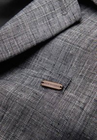 Tissu gris texturé avec une petite étiquette métallique rectangulaire portant la mention « QUINTESSENCE » attachée près d’une couture.