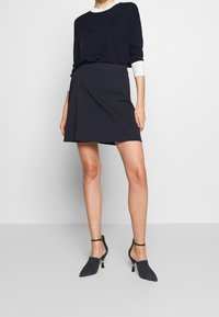 Femme portant un haut à manches longues noir avec des poignets blancs, une jupe portefeuille noire et des chaussures à talons noires avec des brides à la cheville, debout.