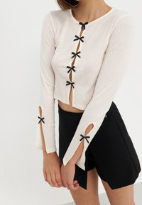 Cremefarbener, gerippter Langarm-Top mit Knopfverschluss und schwarzen Schleifchen-Akzenten, ausgestattet mit Ausschnitten an den Handgelenken und kombiniert mit schwarzen Shorts.