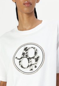 White cotton t-shirt featuring a round logo with black and grey animal print, bordered by silver studs. Short sleeves, loose fit.