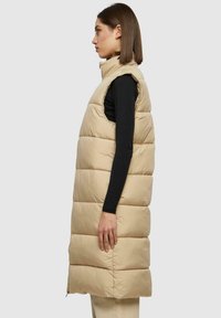 Beige gewatteerde vest, mouwloos met gestikte secties. Ritssluiting aan de voorkant, hoge kraag, lichtgewicht stof en zijsplitten voor bewegingsvrijheid.