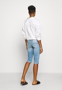 Camisa blanca de gran tamaño, con botones y mangas tres cuartos, combinada con pantalones cortos de mezclilla azules claros, de hasta la rodilla, con aberturas laterales. Sandalias negras tipo slide.