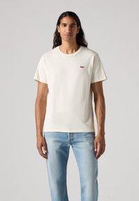 Korte mouwen crèmekleurig T-shirt van katoen met een ronde hals. Bevat een klein rood logo op de borst. G gedragen met lichtblauwe jeans.