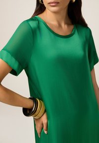 Tunika verde a maniche corte con scollo rotondo, realizzata in tessuto morbido. Accentuata da braccialetti impilati in nero e giallo.