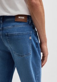 Blå denimjeans med en strukturerad yta. Har en brun läderetikett med "BOSS" och en silverfärgad logotyp på bakfickan.