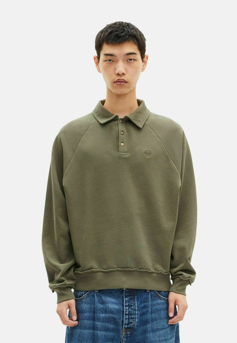Jonge man in een olijfgroene langemouwen polo sweatshirt en een blauwe denim jeans, staand tegen een effen lichte achtergrond.