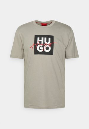 Beige kurzärmliges T-Shirt mit schwarzem quadratischem Logo und fettem weißen "HUGO" Text, überlagert von roter kursiver "Hugo" Schrift.