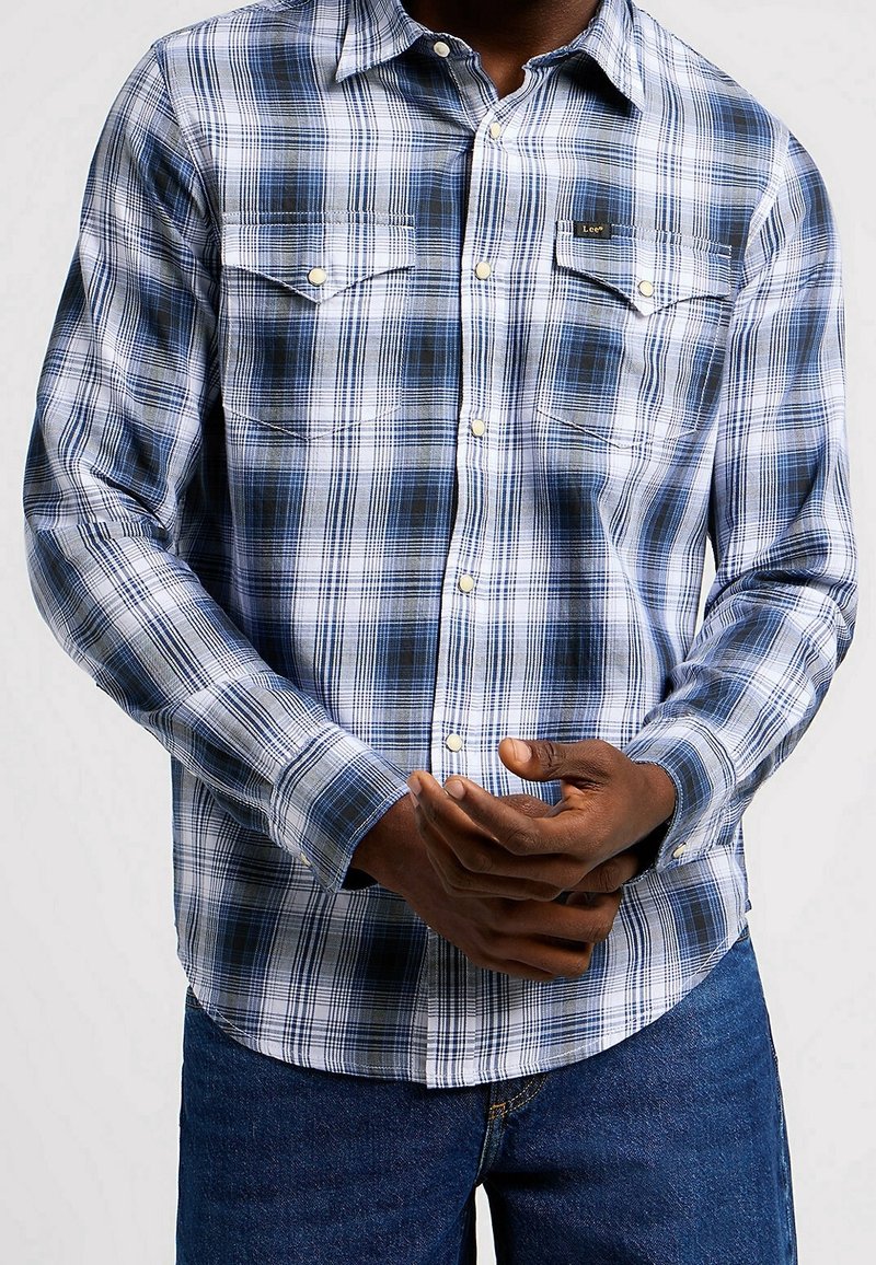 Homme portant une chemise à carreaux bleue et blanche boutonnée avec poches poitrine et un jean bleu, mains jointes devant.