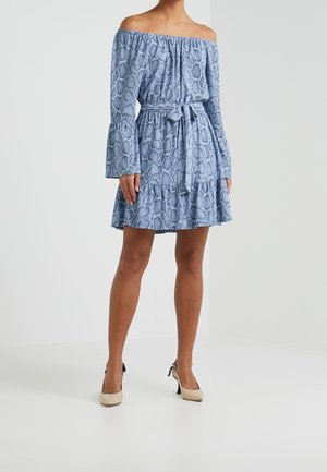 Femme portant une robe bleu épaules dénudées avec imprimé serpent, manches longues évasées, ceinture nouée à la taille, et talons hauts beiges sur fond uni.