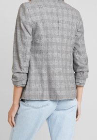 Grårutigt blazerjacka gjort av texturerat tyg, med en strukturerad design, uppvikta ärmar och en ren baksida utan synliga knäppningar.