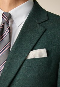 Sacou verde închis din lână cu rever înclinat, asortat cu o cămașă albă și o cravată cu un model în nuanțe de roz și gri. Batista albă este vizibilă.
