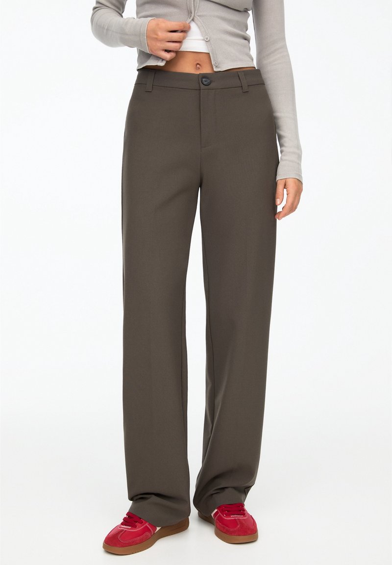 Femme portant un pantalon marron droit, des baskets rouges et un cardigan court gris, debout devant un fond blanc uni.