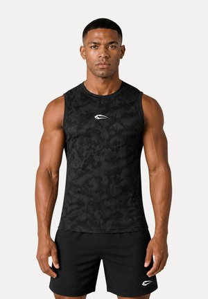 Muskelbåret mand iført en tætsiddende sort camouflage ærmeløs skjorte og sorte shorts, der står med fronten mod en ensfarvet baggrund.